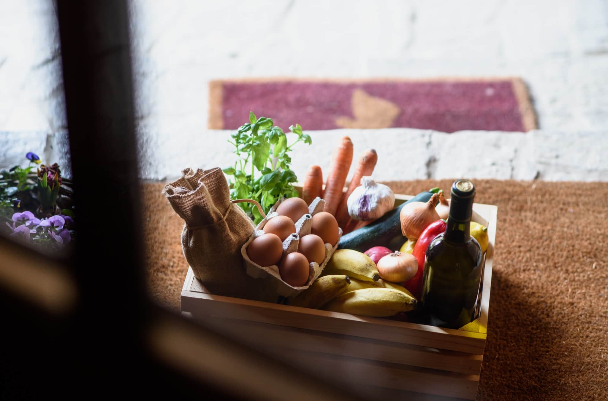 A wooden crate of fresh groceries — eggs, vegetables, fruit, garlic and a bottle of wine — left on a villa doorstep in morning light — grocery delivery on Rhodes